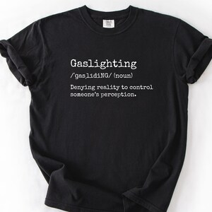 May include: Black t-shirt with the word "Gaslighting" in white, along with its phonetic pronunciation and definition: "Denying reality to control someone's perception."