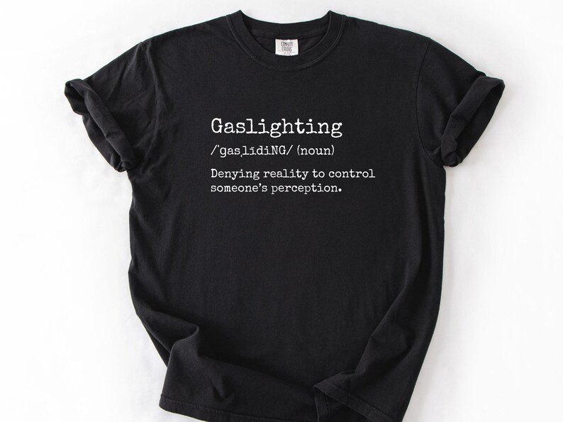 May include: Black t-shirt with the word "Gaslighting" in white, along with its phonetic pronunciation and definition: "Denying reality to control someone's perception."