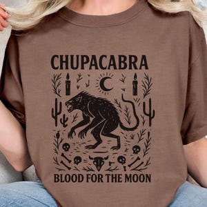May include: Brown t-shirt featuring a black graphic of a Chupacabra, a mythical creature, surrounded by cacti, candles, and a crescent moon. The text "CHUPACABRA" and "BLOOD FOR THE MOON" are printed above and below the graphic.