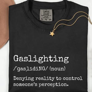May include: Black t-shirt with the word "Gaslighting" in white, along with its definition. A gold necklace with a star pendant rests on the shirt. A brown hat and flowers are in the background.