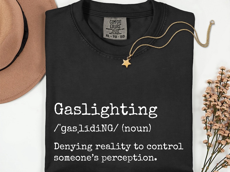 May include: Black t-shirt with the word "Gaslighting" in white, along with its definition. A gold necklace with a star pendant rests on the shirt. A brown hat and flowers are in the background.
