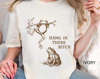 Op de afbeelding: Ivoor kleurig T-shirt met een sepia illustratie van twee kikkers. Eén kikker hangt aan een tak, de andere zit eronder. De tekst "HANG IN THERE BITCH" is vetgedrukt. Het shirt heeft een relaxte pasvorm.