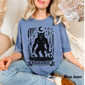 May include: Blue jean colored t-shirt featuring a black Rougarou graphic. The design includes a werewolf, crescent moon, stars, candles, and the word "ROUGAROU" on a banner. The shirt is a comfortable, casual style.