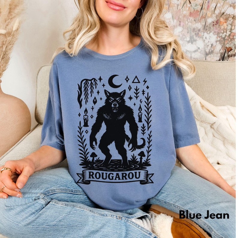 May include: Blue jean colored t-shirt featuring a black Rougarou graphic. The design includes a werewolf, crescent moon, stars, candles, and the word "ROUGAROU" on a banner. The shirt is a comfortable, casual style.