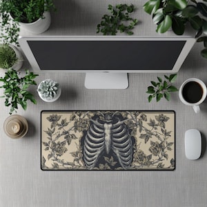Puede incluir: Una alfombrilla de ratón rectangular con una ilustración detallada de una caja torácica humana rodeada de rosas y vides. El diseño es en tonos negros y beige, creando un tema gótico o anatómico. La alfombrilla está sobre una superficie gris.