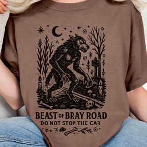 Op de afbeelding: Bruin t-shirt met een zwarte illustratie van een weerwolf, een halve maan, sterren en de tekst "BEAST OF BRAY ROAD DO NOT STOP THE CAR". Het ontwerp bevat bomen, kaarsen en een schedel.