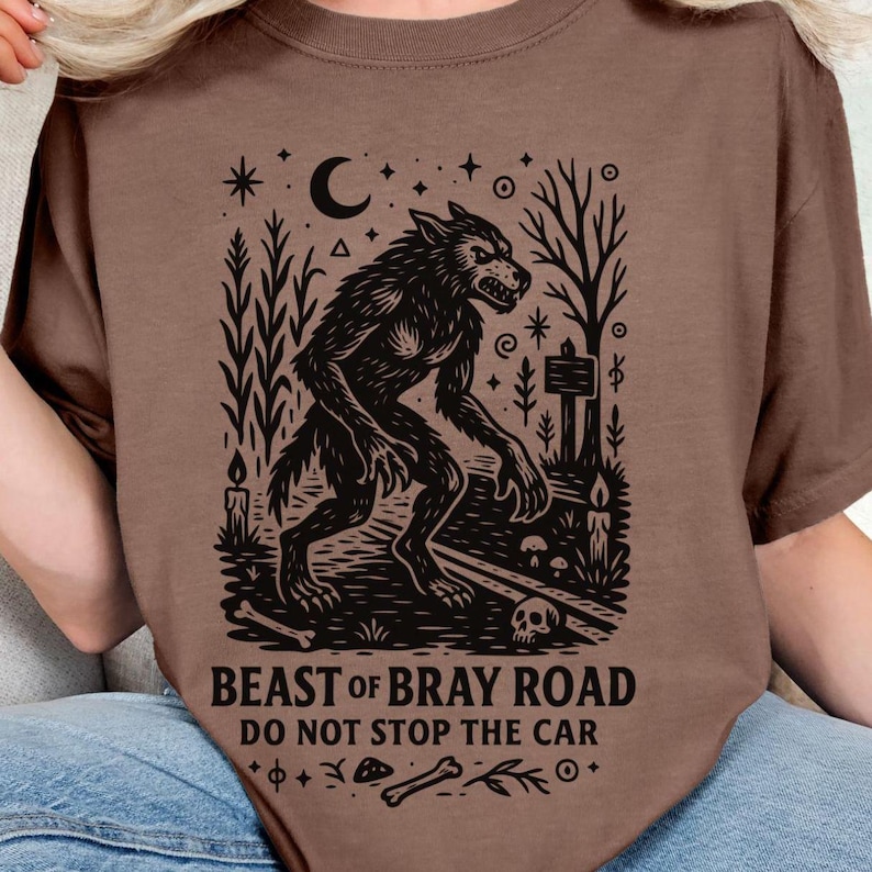 Op de afbeelding: Bruin t-shirt met een zwarte illustratie van een weerwolf, een halve maan, sterren en de tekst "BEAST OF BRAY ROAD DO NOT STOP THE CAR". Het ontwerp bevat bomen, kaarsen en een schedel.
