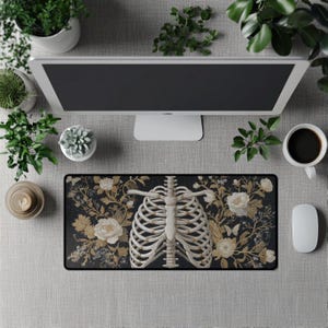 Puede incluir: Alfombrilla de ratón rectangular negra con un diseño de caja torácica y detalles florales blancos y dorados. Un monitor de ordenador, una taza de café, un ratón y plantas rodean la alfombrilla.