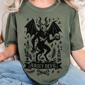 May include: Olive green t-shirt featuring a black graphic of a horned creature with bat wings holding a skull. The text "JERSEY DEVIL" is displayed on a banner below the creature, with crossbones and stars.