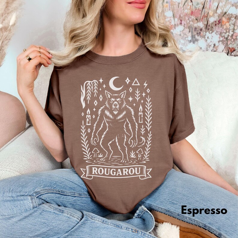 May include: Espresso-colored t-shirt featuring a white illustration of a werewolf with a crescent moon, geometric shapes, and the word "ROUGAROU" on a banner. The shirt is a solid color.