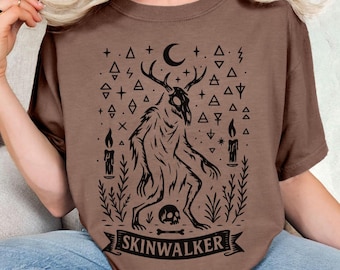 May include: Brown t-shirt featuring a black graphic of a creature with antlers, a skull, and the word "SKINWALKER" on a banner. The design includes a crescent moon, candles, and geometric shapes. The shirt is a solid brown color.