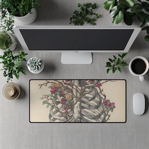 Puede incluir: Alfombrilla de ratón rectangular con una ilustración detallada de una caja torácica humana entrelazada con vides florecientes. La obra de arte está en tonos grises y beige, con flores coloridas. La alfombrilla tiene un borde negro y está rodeada de material de oficina.