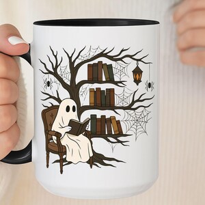 Op de afbeelding: Witte keramische mok met een zwarte binnenkant en handvat. De mok heeft een grillige illustratie van een spook dat een boek leest in een stoel, met een boom, boeken, spinnen en een lantaarn. Het ontwerp heeft een Halloween-thema.