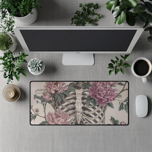 Puede incluir: Alfombrilla de ratón rectangular con una ilustración detallada de una caja torácica humana entrelazada con peonías rosas y hojas verdes. El diseño está sobre un fondo beige con un borde negro. También se ven un monitor de ordenador, una taza de café y un ratón.