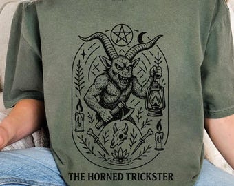 Könnte beinhalten: Olivgrünes T-Shirt mit einer schwarzen Grafik eines gehörnten Wesens, das eine Axt und eine Laterne hält, umgeben von Kerzen, Messern und einem Pentagramm. Der Text "THE HORNED TRICKSTER" ist darunter gedruckt.