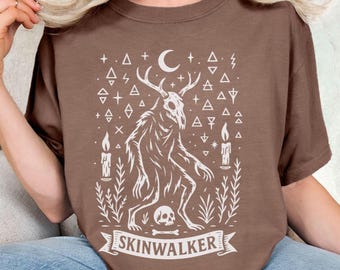 May include: Brown t-shirt featuring a white graphic of a creature with antlers, a skull, and a banner that reads "SKINWALKER." The design includes candles, a crescent moon, and geometric shapes. The shirt is made of a soft material.