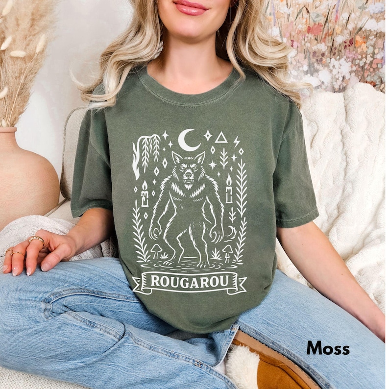 May include: Moss green t-shirt featuring a white illustration of a werewolf with a crescent moon, stars, and the word "ROUGAROU" on a banner. The shirt is a relaxed fit.