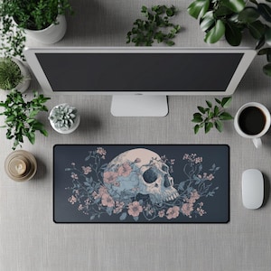 Puede incluir: Alfombrilla de ratón rectangular con una ilustración detallada de una calavera adornada con flores azules y rosas. El diseño está sobre un fondo gris oscuro. La alfombrilla tiene un borde negro.