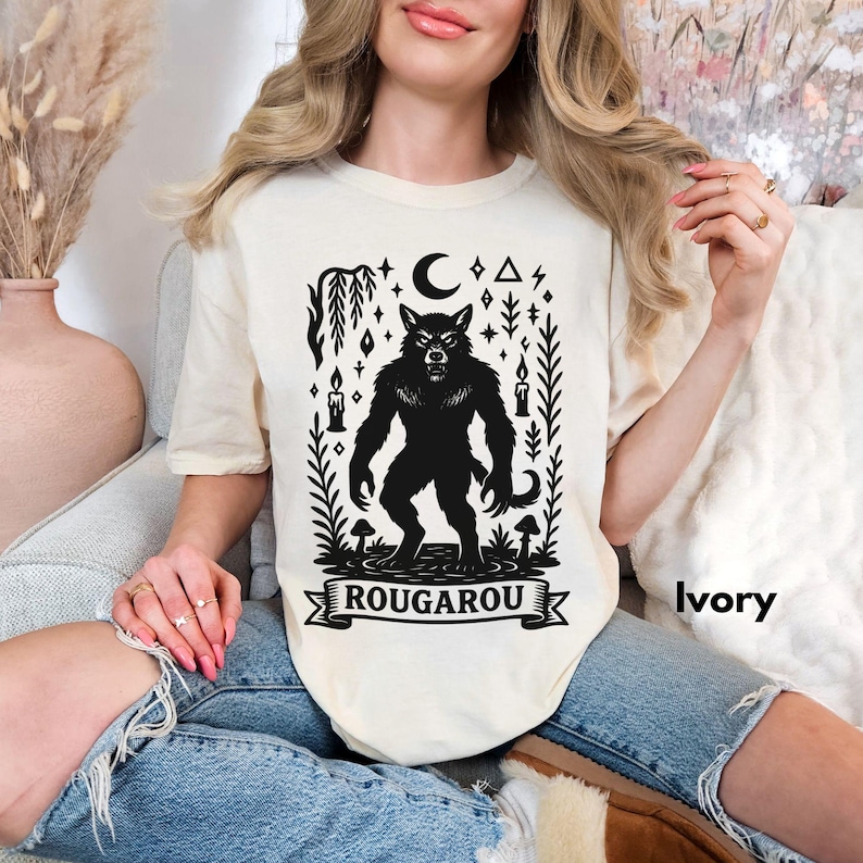 May include: Ivory-colored t-shirt featuring a black graphic of a werewolf with a crescent moon, stars, and candles. The word "ROUGAROU" is printed on a banner below the werewolf. The shirt is made of a soft material.