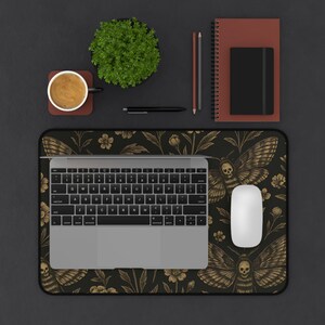 May include: A black and gold mouse mat featuring a moth and floral pattern, a laptop, and a white mouse. The desk also has a coffee cup, a small plant, a pen, pencils, and a notebook.