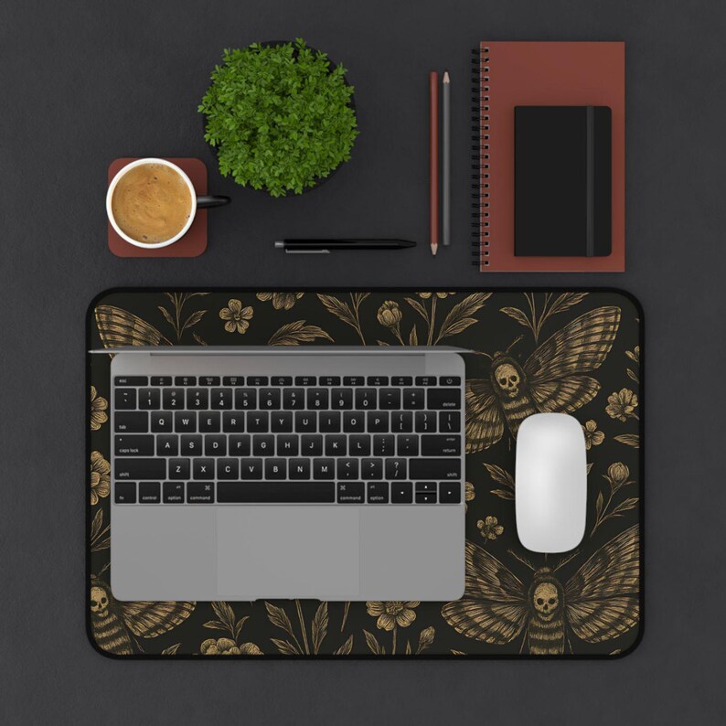 May include: A black and gold mouse mat featuring a moth and floral pattern, a laptop, and a white mouse. The desk also has a coffee cup, a small plant, a pen, pencils, and a notebook.