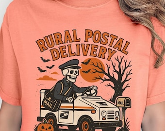 May include: Coral-colored t-shirt featuring a Halloween-themed graphic. The design includes a skeleton driving a white mail truck with the text "RURAL POSTAL DELIVERY". A jack-o'-lantern, bats, and a tree are also included in the design.