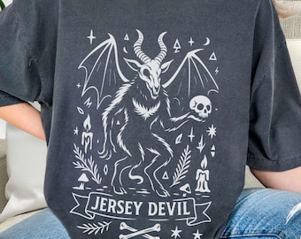May include: Dark gray t-shirt featuring a white graphic of the Jersey Devil, a mythical creature with horns, wings, and a skull. The text "JERSEY DEVIL" is displayed on a banner below the illustration, with bones and stars.