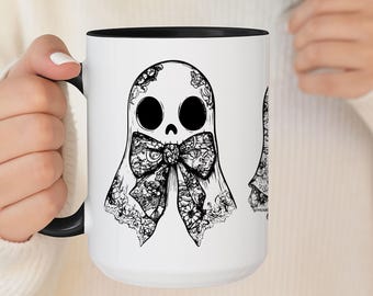 May include: White ceramic mug with a black interior and handle. The mug features a black and white illustration of a ghost with large eyes and a floral bow. The ghost design is repeated on the mug.