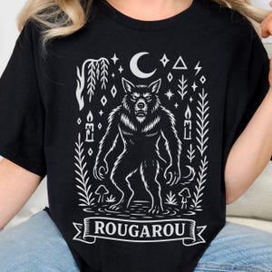 May include: Black t-shirt featuring a white illustration of a werewolf, a crescent moon, stars, and foliage. The word "ROUGAROU" is displayed in a banner at the bottom of the design. The graphic is centered on the shirt.