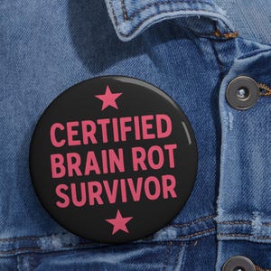 Puede incluir: Un botón negro con una estrella rosa a cada lado y el texto "CERTIFIED BRAIN ROT SURVIVOR" en rosa.