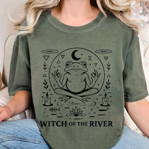 May include: Olive green t-shirt featuring a black graphic of a meditating frog inside a circle with a crescent moon, candles, and a lotus flower. The text "WITCH OF THE RIVER" is printed below the graphic.