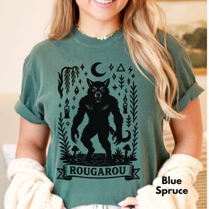 May include: Blue Spruce t-shirt featuring a black Rougarou graphic. The design includes a werewolf, a crescent moon, candles, and the word "ROUGAROU" on a banner. The shirt is a solid color.