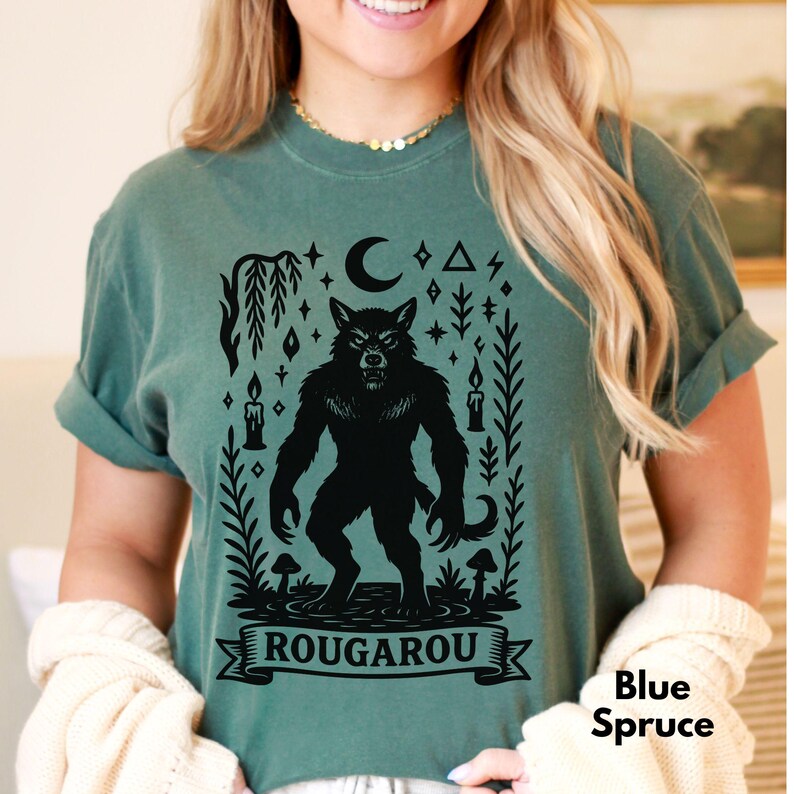 May include: Blue Spruce t-shirt featuring a black Rougarou graphic. The design includes a werewolf, a crescent moon, candles, and the word "ROUGAROU" on a banner. The shirt is a solid color.