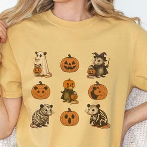 K&ouml;nnte beinhalten: Senfgelbes T-Shirt mit Halloween-Illustrationen von Opossums und K&uuml;rbissen. Die Opossums sind als Geist, Hexe, Mumie und Skelett verkleidet und halten K&uuml;rbislaternen. Die K&uuml;rbisse haben geschnitzte Gesichter. Ein lustiges und festliches Design f&uuml;r Halloween.