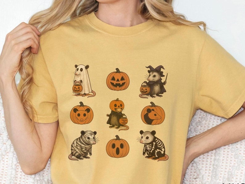 K&ouml;nnte beinhalten: Senfgelbes T-Shirt mit Halloween-Illustrationen von Opossums und K&uuml;rbissen. Die Opossums sind als Geist, Hexe, Mumie und Skelett verkleidet und halten K&uuml;rbislaternen. Die K&uuml;rbisse haben geschnitzte Gesichter. Ein lustiges und festliches Design f&uuml;r Halloween.