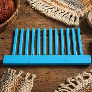 May include: A blue weaving tool with vertical bars is the focal point of this image, surrounded by craft supplies. A basket of yarn, wooden beads, and a shuttle are also visible. The items are arranged on a wooden surface, suggesting a crafting theme.