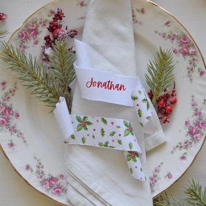 Pode incluir: Uma toalha de linho branca sobre um prato floral com borda dourada, adornada com uma fita que diz "Jonathan" em escrita vermelha. A fita é decorada com um padrão de azevinho e bagas. Decoração festiva de férias.