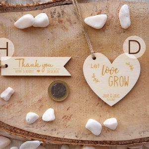 K&ouml;nnte beinhalten: Zwei Holz-Hochzeitsgeschenke auf einem Holz-Hintergrund. Ein Geschenk ist ein herzf&ouml;rmiges Schild mit dem Text "Let love grow" und den Namen "Julie & Louis" sowie dem Datum "12.12.2021". Das andere Geschenk ist ein rechteckiges Schild mit dem Text "Thank you" mit einem Herz und dem Text "Julie & Louis 12.12.2021".