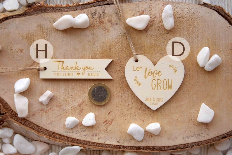 K&ouml;nnte beinhalten: Zwei Holz-Hochzeitsgeschenke auf einem Holz-Hintergrund. Ein Geschenk ist ein herzf&ouml;rmiges Schild mit dem Text "Let love grow" und den Namen "Julie & Louis" sowie dem Datum "12.12.2021". Das andere Geschenk ist ein rechteckiges Schild mit dem Text "Thank you" mit einem Herz und dem Text "Julie & Louis 12.12.2021".