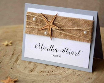 Peut inclure: Une carte de couleur gris clair avec un ruban en toile de jute et une étoile de mer en bois. La carte indique "Martha Stuart, Table 4".