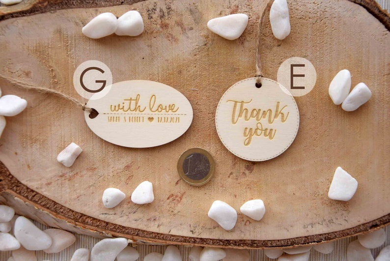 K&ouml;nnte beinhalten: Zwei Holz-Geschenk-Etiketten mit goldener Beschriftung. Eine Etikette ist oval geformt und sagt "with love" mit einem Herzen und Namen. Die andere Etikette ist rund und sagt "Thank you".