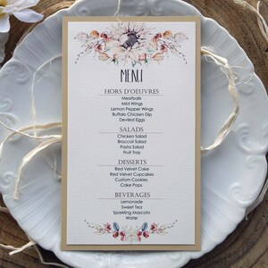 Pode incluir: Um cartão de menu bege com um design floral em aquarela. O menu inclui aperitivos, saladas, sobremesas e bebidas. O texto está em preto e azul.