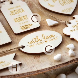 K&ouml;nnte beinhalten: Ein Set aus acht Holz-Hochzeitsgeschenkanh&auml;ngern mit verschiedenen Designs und Texten. Die Anh&auml;nger sind alle wei&szlig; mit goldener Beschriftung. Einige der Anh&auml;nger haben eine Herzform, w&auml;hrend andere rechteckig oder oval sind. Der Text auf den Anh&auml;ngern lautet "Danke", "Mit Liebe", "Ein s&uuml;&szlig;er Abschluss f&uuml;r einen neuen Anfang" und "Mit Liebe J&R".