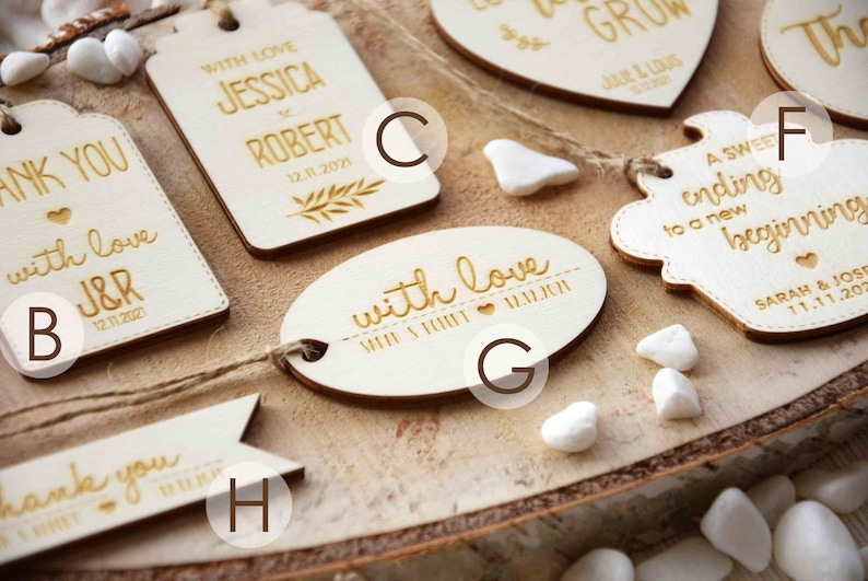 K&ouml;nnte beinhalten: Ein Set aus acht Holz-Hochzeitsgeschenkanh&auml;ngern mit verschiedenen Designs und Texten. Die Anh&auml;nger sind alle wei&szlig; mit goldener Beschriftung. Einige der Anh&auml;nger haben eine Herzform, w&auml;hrend andere rechteckig oder oval sind. Der Text auf den Anh&auml;ngern lautet "Danke", "Mit Liebe", "Ein s&uuml;&szlig;er Abschluss f&uuml;r einen neuen Anfang" und "Mit Liebe J&R".