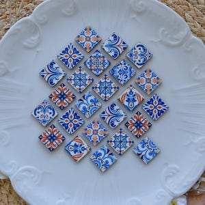Puede incluir: Una colección de 18 pequeños azulejos cuadrados de cerámica con intrincados patrones azules, blancos y naranjas. Los azulejos están dispuestos en un patrón circular sobre un plato blanco.