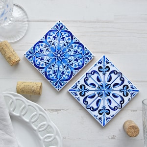 May include: Two blue and white ceramic tile coasters with intricate floral patterns. The coasters are on a white wooden surface.