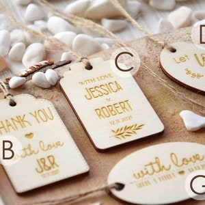 K&ouml;nnte beinhalten: Sortiment aus Holz-Hochzeitsgeschenkanh&auml;ngern mit goldener Beschriftung. Die Anh&auml;nger zeigen verschiedene Designs, darunter "Danke", "Mit Liebe", "Lass die Liebe wachsen" und "Jessica & Robert".