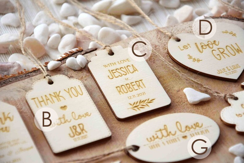 K&ouml;nnte beinhalten: Sortiment aus Holz-Hochzeitsgeschenkanh&auml;ngern mit goldener Beschriftung. Die Anh&auml;nger zeigen verschiedene Designs, darunter "Danke", "Mit Liebe", "Lass die Liebe wachsen" und "Jessica & Robert".