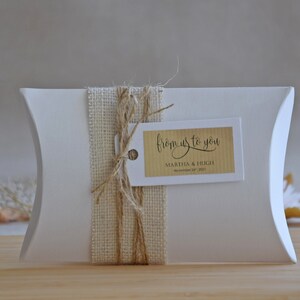 May include: White pillow box with a burlap ribbon and twine bow. A brown tag with the text "from us to you" is attached to the bow. The tag also includes the names "Martha & Hugh" and the date "November 19, 2023".