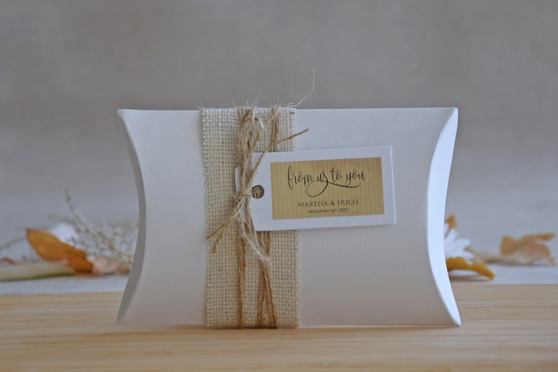 May include: White pillow box with a burlap ribbon and twine bow. A brown tag with the text "from us to you" is attached to the bow. The tag also includes the names "Martha & Hugh" and the date "November 19, 2023".